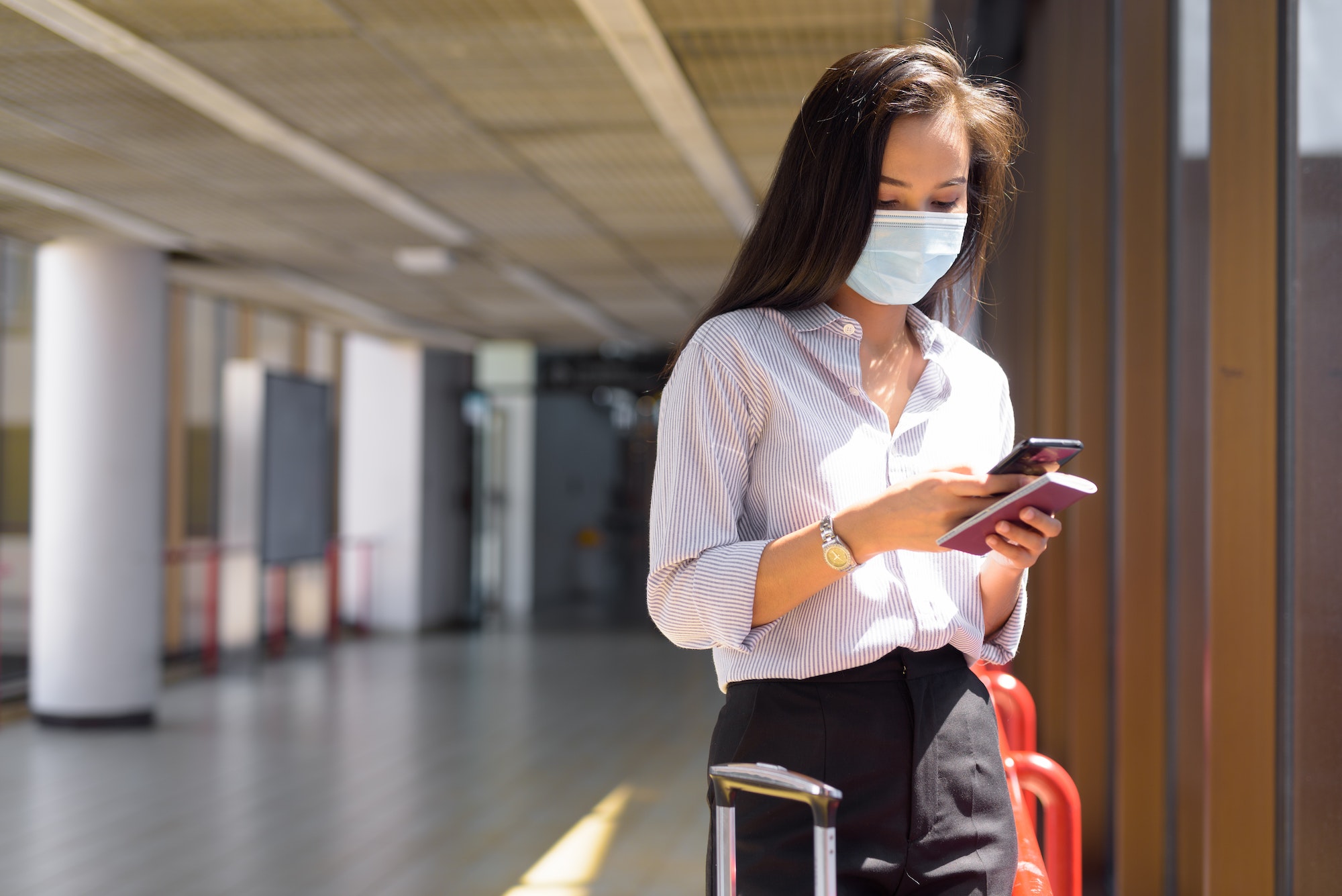 Young Asian tourist woman with mask using phone and holding passport at the airport